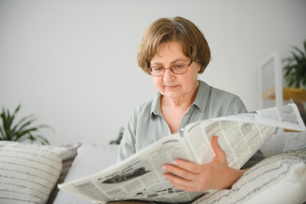 Senior lady reading her newspaper at home relaxing on a couch and peering over the top at the viewer