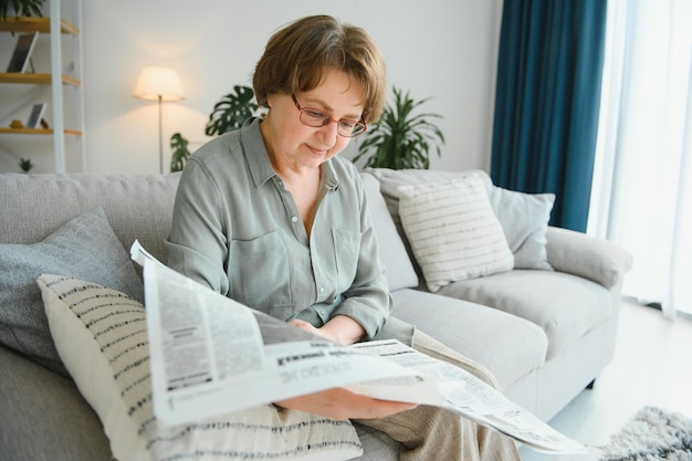 Пожилая дама читает газету дома, отдыхает на диване и смотрит сверху на зрителя