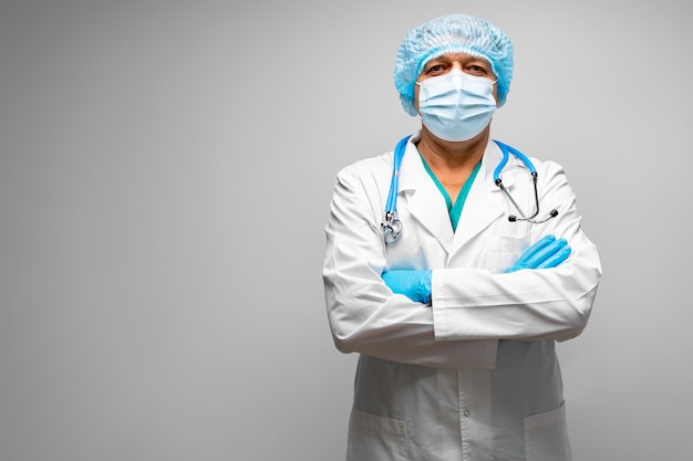 Senior male doctor in face mask standing against grey background