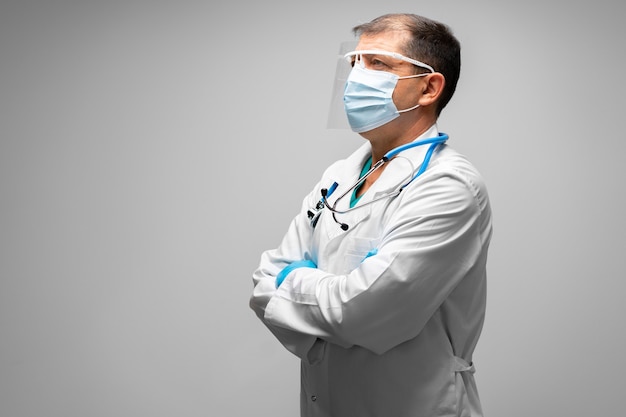 Senior male doctor in face mask standing against grey portrait