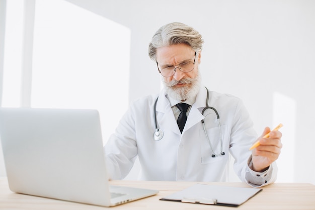 Senior male Doctor is thinking and checking his patient medical records on a laptop computer in the medical room