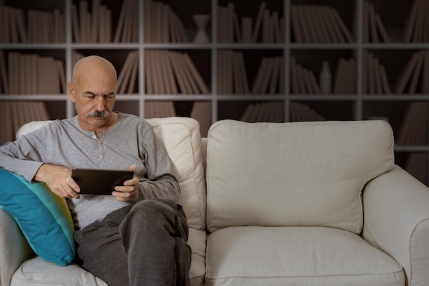 Uomo anziano che legge in una tavoletta digitale seduto sul divano del soggiorno di casa