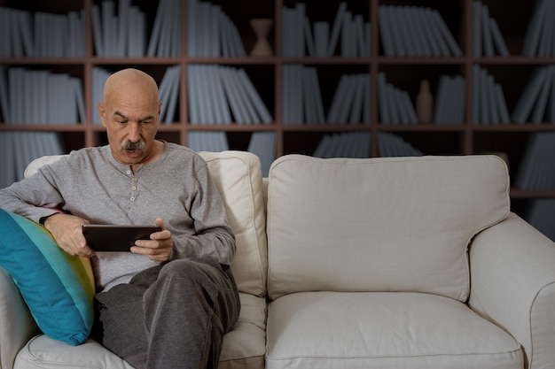 Senior male reading in a digital tablet sitting at the sofa at\
the house living room