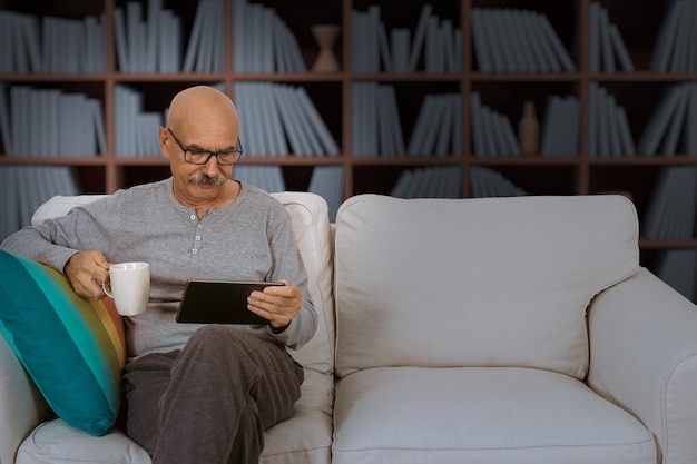 Senior Male Reading in a Digital Tablet Sitting at the Sofa at the House Living Room