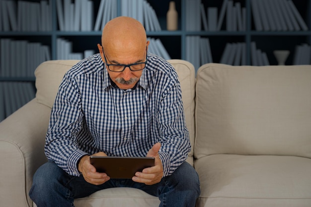 家のリビングルームのソファに座っているデジタルタブレットで読書をしている年配の男性