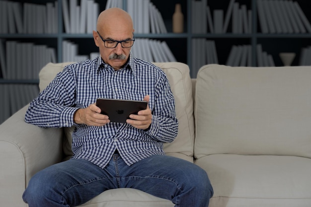 Senior male reading in a digital tablet sitting at the sofa at\
the house living room