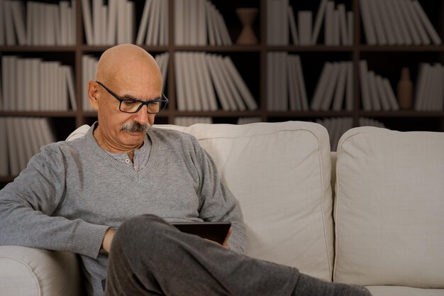 Старший мужчина читает на цифровом планшете, сидя на диване в гостиной дома