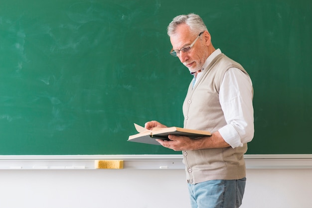 senior male teacher reading book against blackboard
