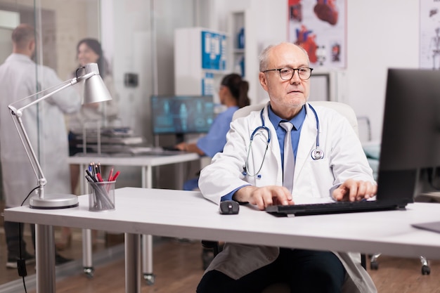 Senior man arts met grijs haar die op de computer werkt terwijl een jonge collega met een patiënt in de ziekenhuisgang is en een verpleegster die het analyserapport controleert.