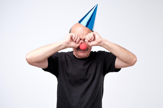 誕生日の帽子をかぶった年配の男性がパーティーで泣いて涙を拭く