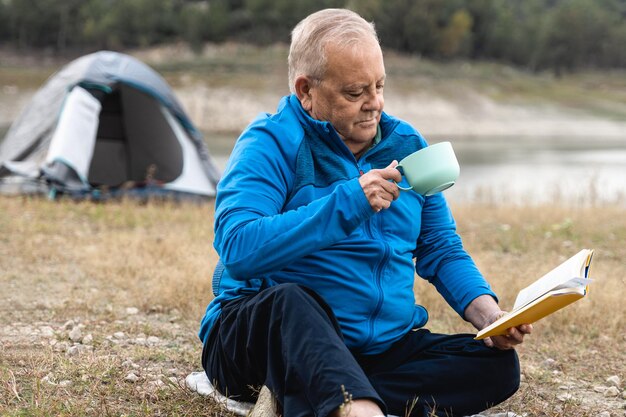 Senior man camping with tent while reading book and drinks\
coffee with lake on background