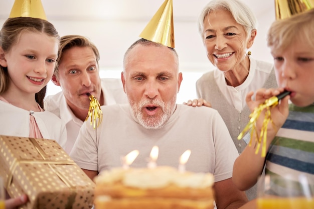 パーティーハットをかぶって笛を吹く家で家族と一緒に誕生日を祝う年配の男性おじいちゃんがバースデーキャンドルを吹き飛ばし、孫の妻と息子に囲まれて願い事をする