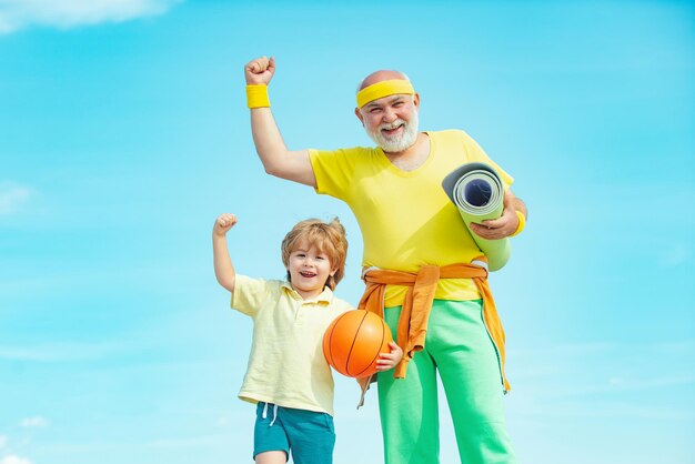 Senior man and child exercising on blue sky Sports for kids Sportsman grandfather and healthy kid with basketball ball on blue sky background