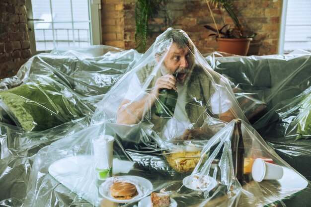 Senior man covered with plastic, eating fast food and drinking beer -