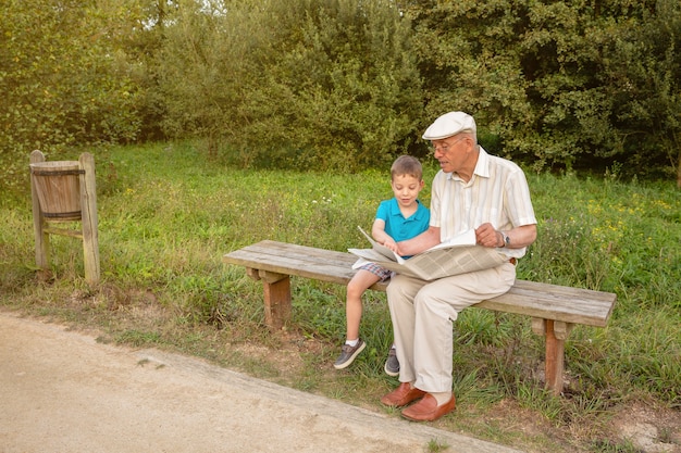 公園のベンチに座って新聞を読んでいる年配の男性とかわいい子供。 2つの異なる世代の概念。