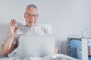 Senior man die op de medische afdeling ligt en met iemand praat via een videogesprek met een laptop