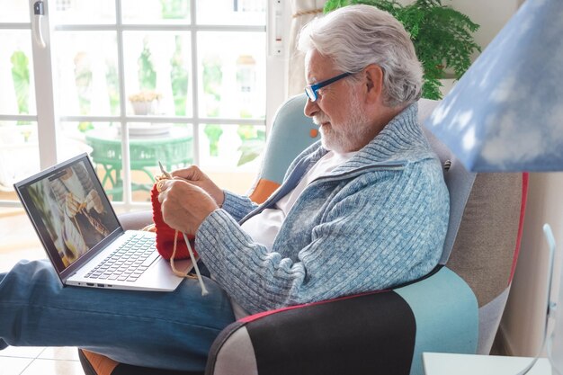 Senior man die thuis leert breien met wollen garen, kijkt naar een online tutorial voor laptops