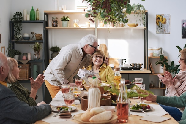 Senior man die vrouw kust en haar feliciteert met vakantie tijdens het diner aan tafel met vrienden