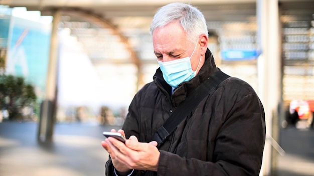 Senior man die zijn smartphone buiten gebruikt terwijl hij een masker draagt ter bescherming tegen de coronavirus-pandemie