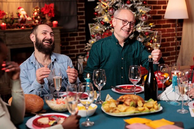 Senior man doet toast met verhoogd glas wijn tijdens het kerstavonddiner om de vakantie van december te vieren met vrienden en familie. Grootvader houdt een toespraak rond de tafel, sprankelende alcohol.
