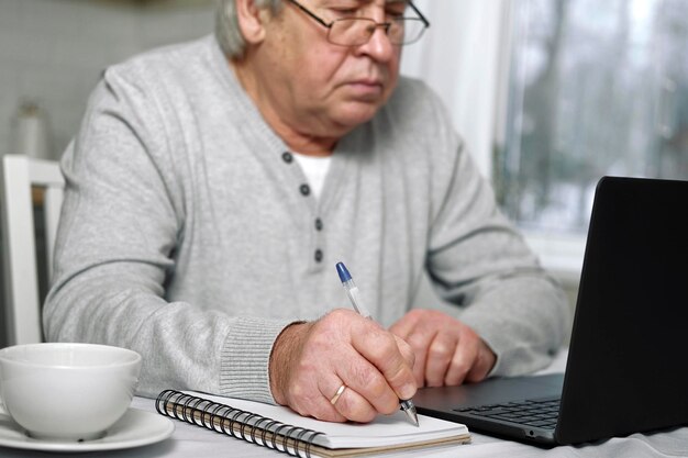 Senior man Elearning schrijven in Kladblok Oude man studeren thuis zitten aan bureau met behulp van laptop Close-up hand schrijft in notebook met pen notitie informatie maken Ouderen afstandsonderwijs concept