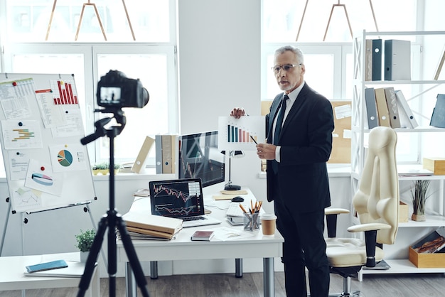 Senior man in elegant business suit pointing at chart while making social media video