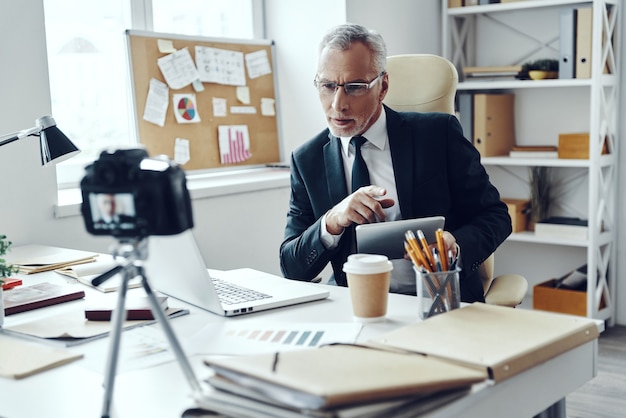 Senior man in elegant business suit using digital tablet while making social media video