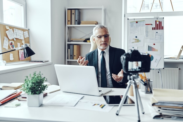 Senior man in elegant shirt and tie working using laptop while making social media video