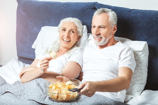 Senior man en vrouw tv kijken en chips eten in het bed en kijken naar de camera