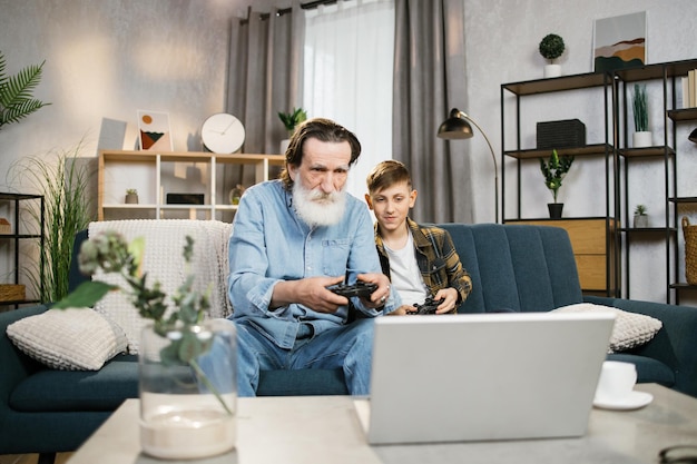 Senior man en zijn kleinzoon in casual kleding met behulp van draadloze joysticks voor het spelen van videogames