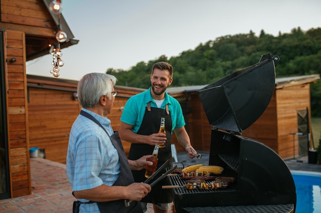 Senior man en zijn zoon genieten in de achtertuin en grillen vlees terwijl ze bier drinken