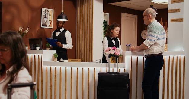 荷物を持ってホテルのロビーに入り、チェックインするコンシェルジュサービスを求める年配の男性。オールインクルーシブのホリデーリゾート、観光業界で年配の観光客にリラックスした滞在を保証する受付係の女性。