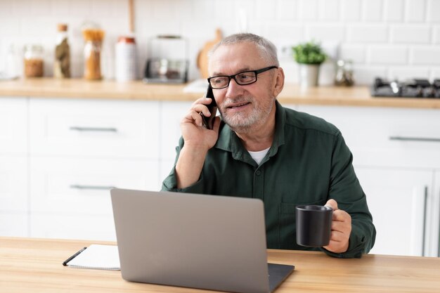 Senior man entrepreneur talking on phone with laptop sitting at the table in the home office