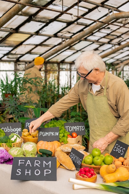 Foto uomo maggiore in occhiali e grembiule in piedi al bancone e preparare alimenti biologici per la vendita al mercato degli agricoltori