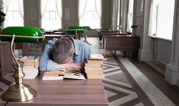 L'uomo anziano si addormenta su un libro di testo è seduto a un tavolo disseminato di materiale didattico