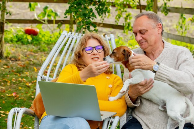 Senior man and female sitting in armchairs with a dog and using laptop in a garden