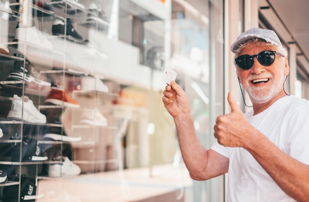 Senior man in front of shop windows holding credit card for payment ready to profit summer sales