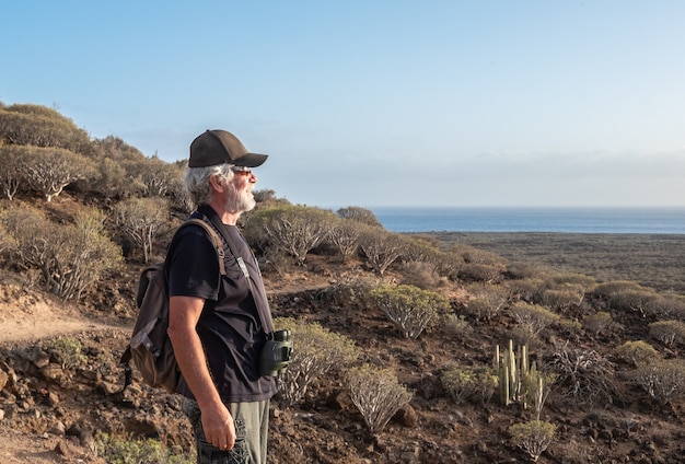 Senior man genieten van wandelen in dorre vulkanische landschap. Kijkend naar de horizon over zee. Rugzak en verrekijker, jeugdige ouderen gezonde levensstijl