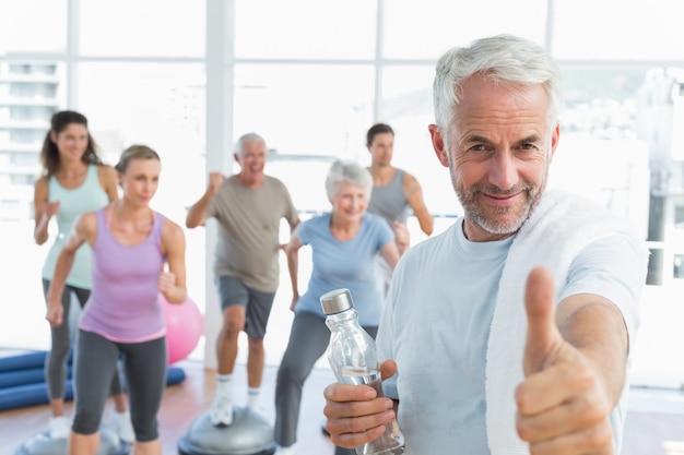 Uomo senior che gesturing i pollici su con la gente che si esercita nello studio di forma fisica