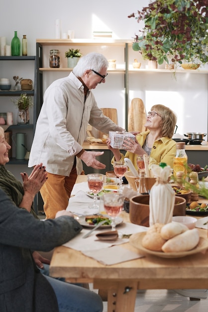 Senior man giving present to woman and congratulating her with birthday while they celebrating at dining table