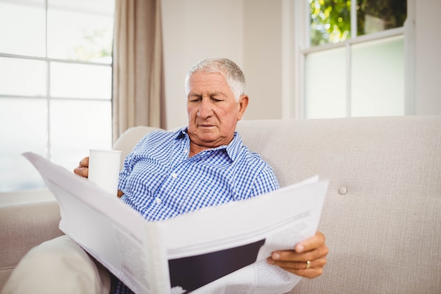 Senior man having a cup of coffee and reading newspaper in living room