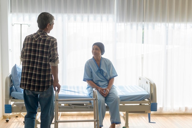 Пожилой мужчина помогает больной раком женщине в платке с ходунками в больнице здравоохранения и медицинской концепции