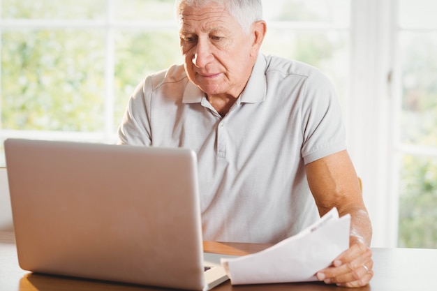 Старший мужчина держит документы и с помощью ноутбука на дому