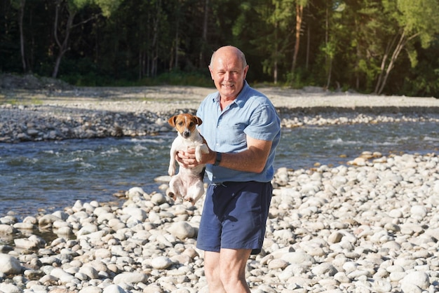 Старший мужчина держит в руке маленькую мокрую собаку Джека Рассела терьера после того, как она купалась в ближайшей реке в солнечный день