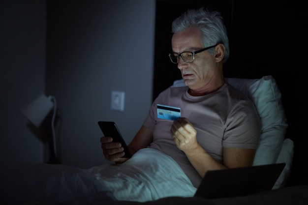 Senior man in bril liggend op bed online betalen met creditcard met zijn mobiele telefoon is het laat in de avond