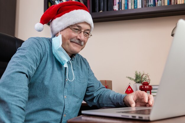 Senior man in kerstman hoed praat met behulp van laptop voor videogesprek vrienden en kinderen. De kamer is feestelijk versierd. Kerst tijdens coronavirus.