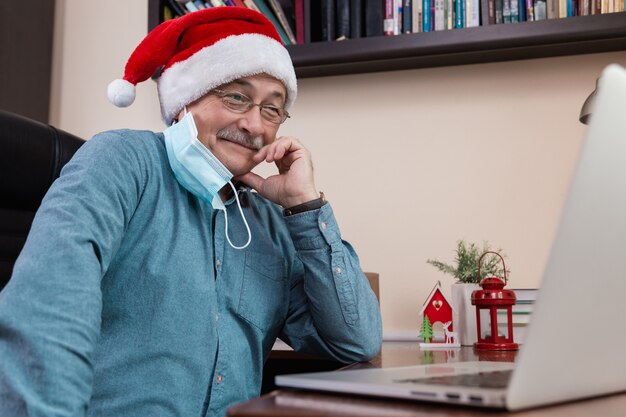 Senior man in kerstman hoed praat met behulp van laptop voor videogesprek vrienden en kinderen. De kamer is feestelijk versierd. Kerst tijdens coronavirus.