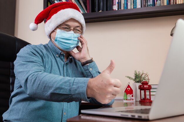 Senior man in kerstman hoed praat met behulp van laptop voor videogesprek vrienden en kinderen. De kamer is feestelijk versierd. Kerst tijdens coronavirus.