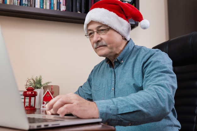 Senior man in kerstman hoed praat met laptop apparaat voor video-oproep vrienden en kinderen. De kamer is feestelijk versierd.