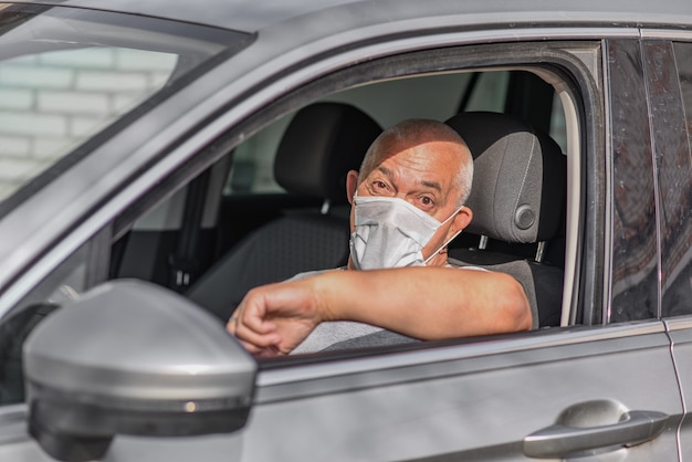 Senior man in medisch masker autorijden, kijkend naar de camera.